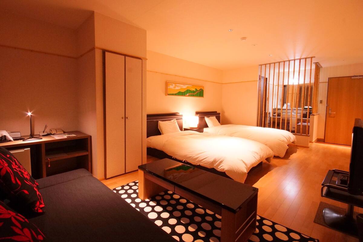 Quarto de hotel com duas camas, uma mesa de trabalho com abajur, televisão e porta de correr.