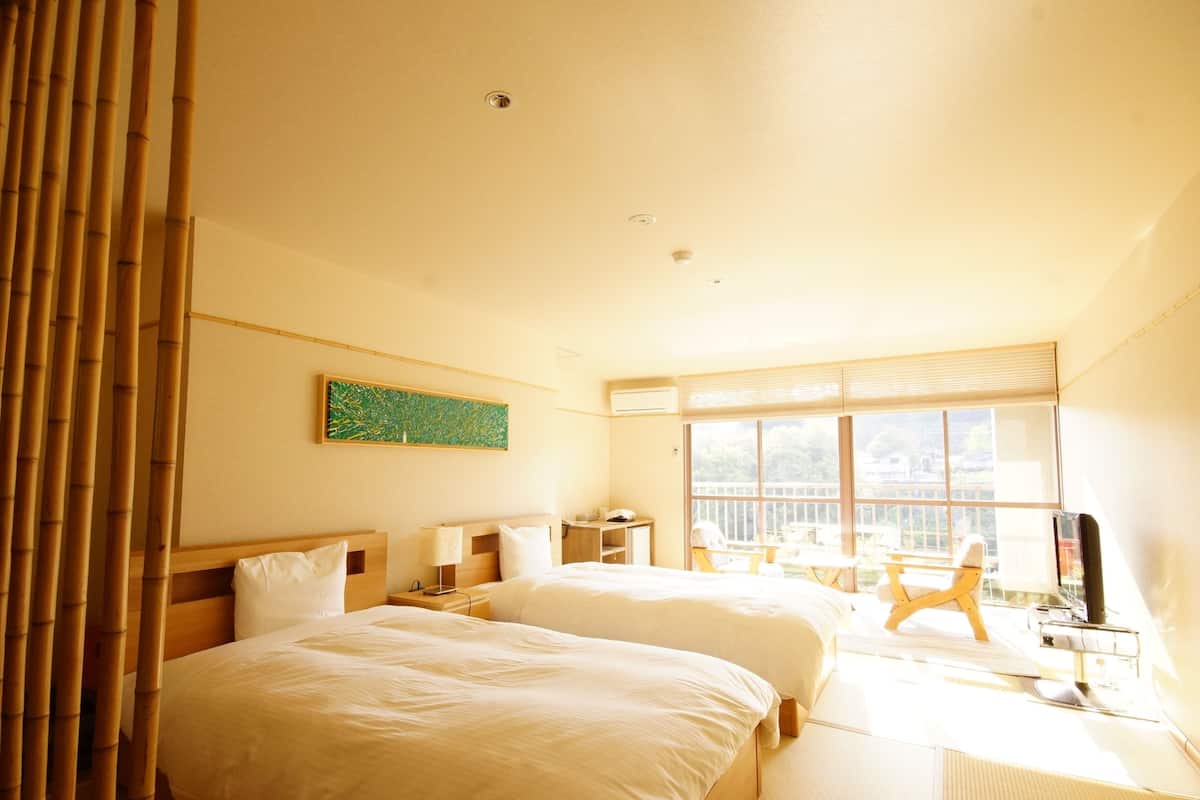 Quarto de hotel com duas camas, uma escrivaninha, uma cadeira, janela com vista e um quadro na parede.
