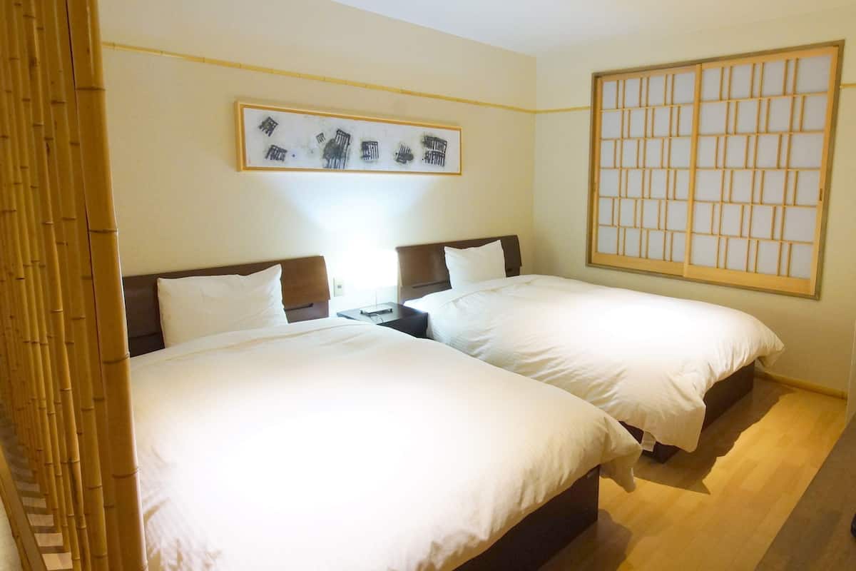 Quarto de hotel com duas camas, uma janela grande com grade de madeira e um quadro na parede.