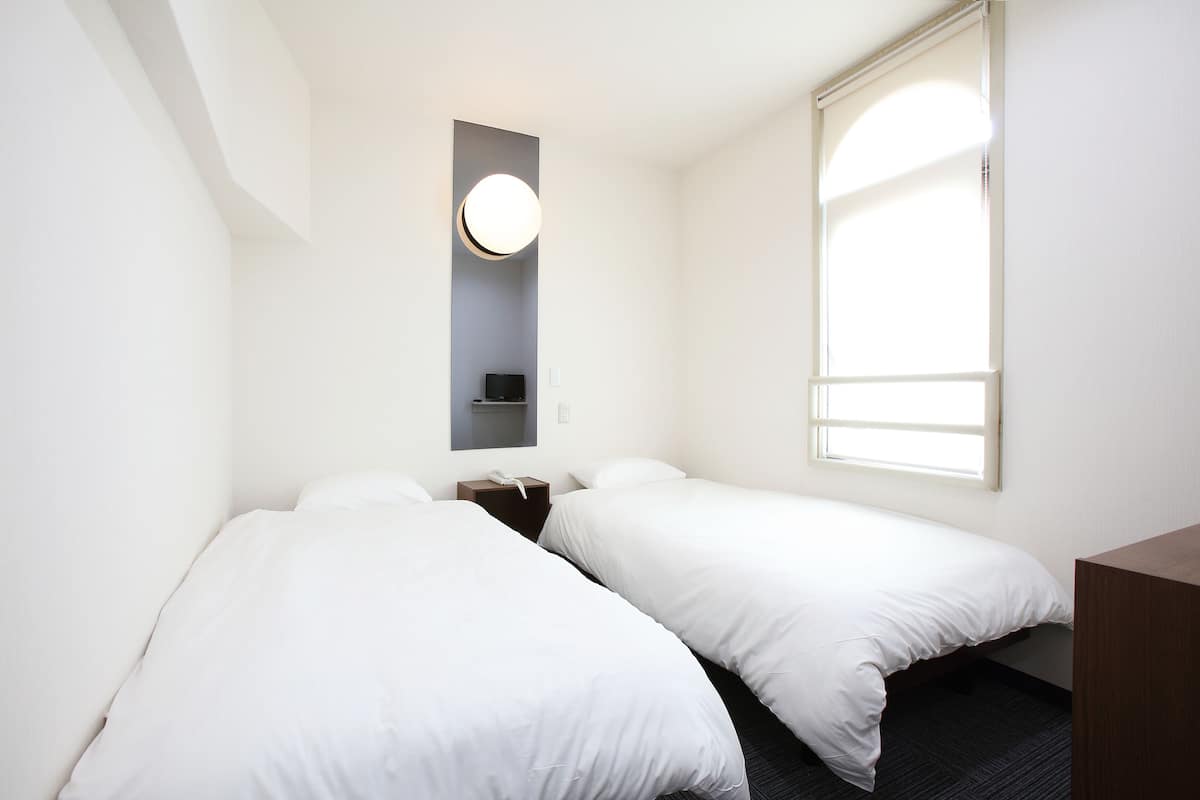 A hotel room with two single beds, a bedside table with a lamp, a small shelf, and a window with blinds.