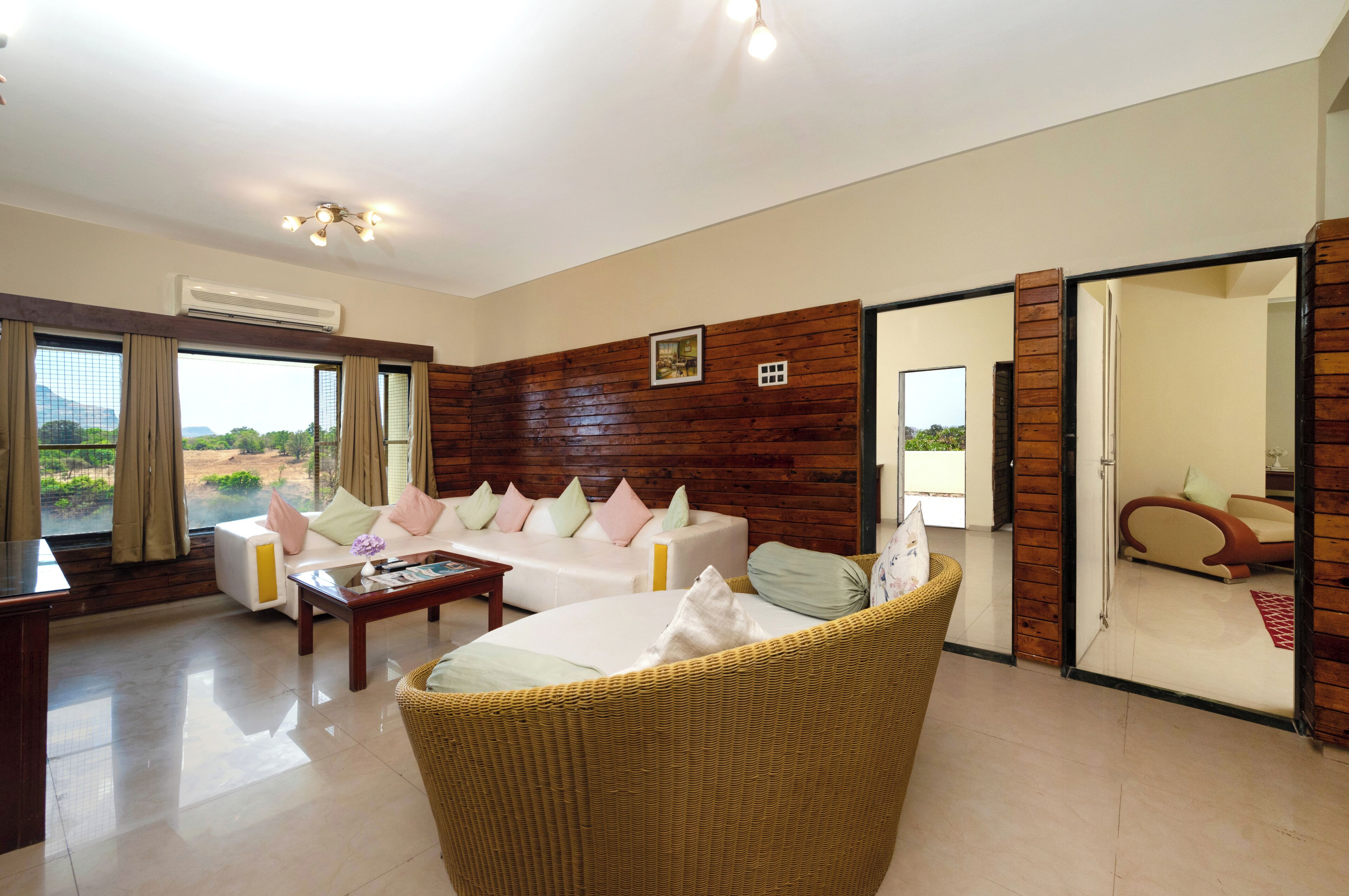 A spacious living room with a sofa set, a wicker chair, and a coffee table. There is a large window with curtains, a wooden wall panel, and a door leading to another room.