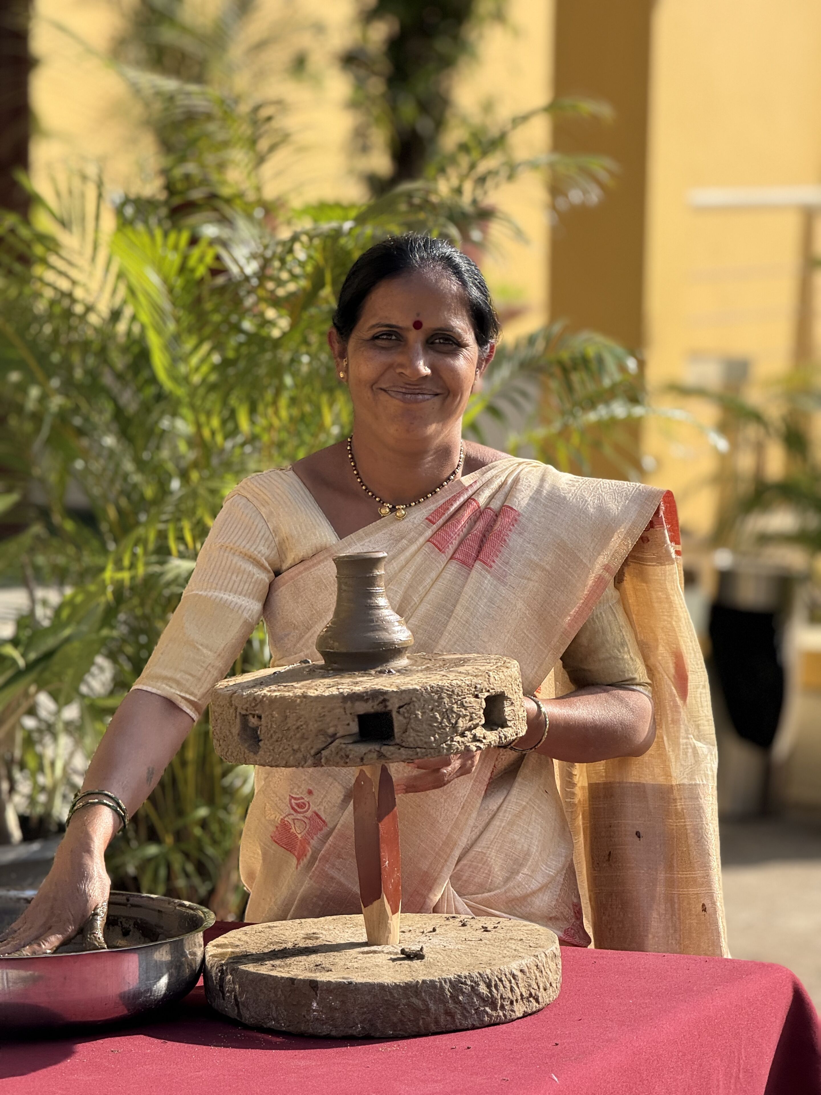 A person in traditional attire holding a clay pot and a metal bowl on a red table.