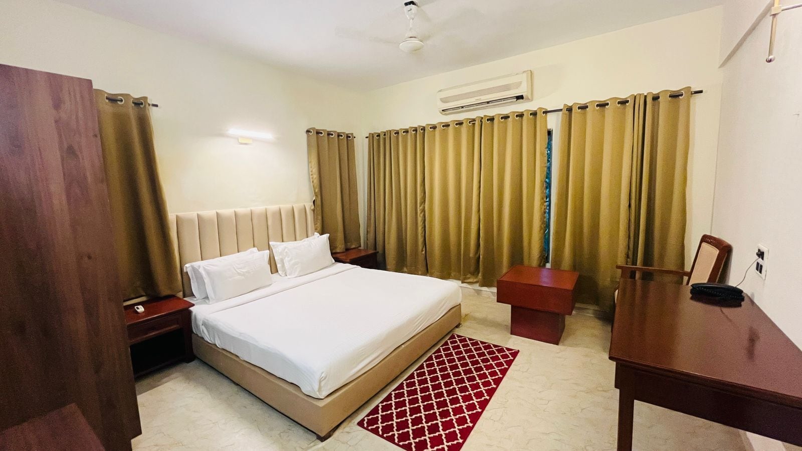 Une chambre d’hôtel avec un lit, un bureau, une chaise, une table de chevet et un tapis rouge à motifs.