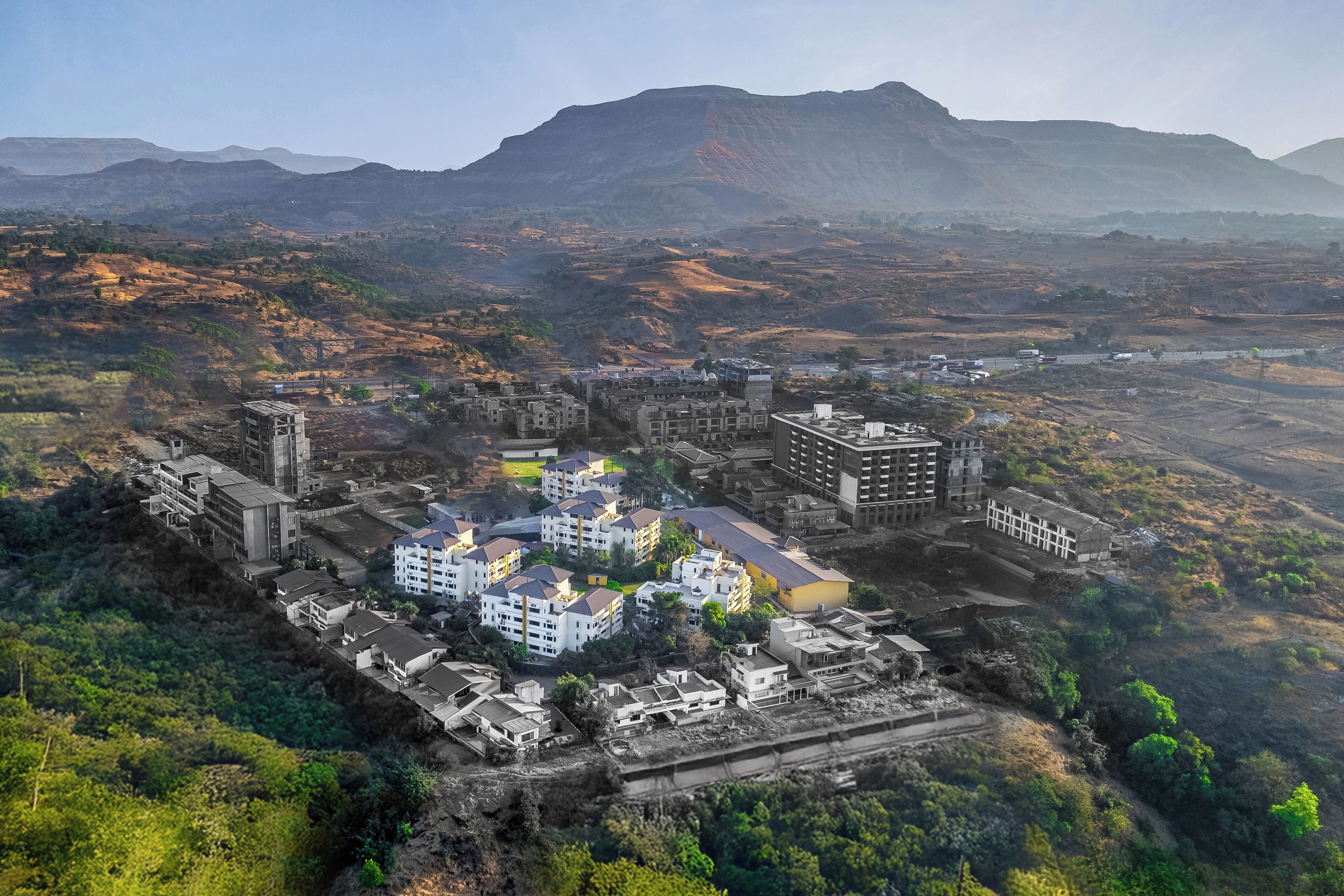 An aerial view of a residential area with multiple buildings, including a mix of apartment complexes and standalone houses, surrounded by greenery and a mountain in the background.