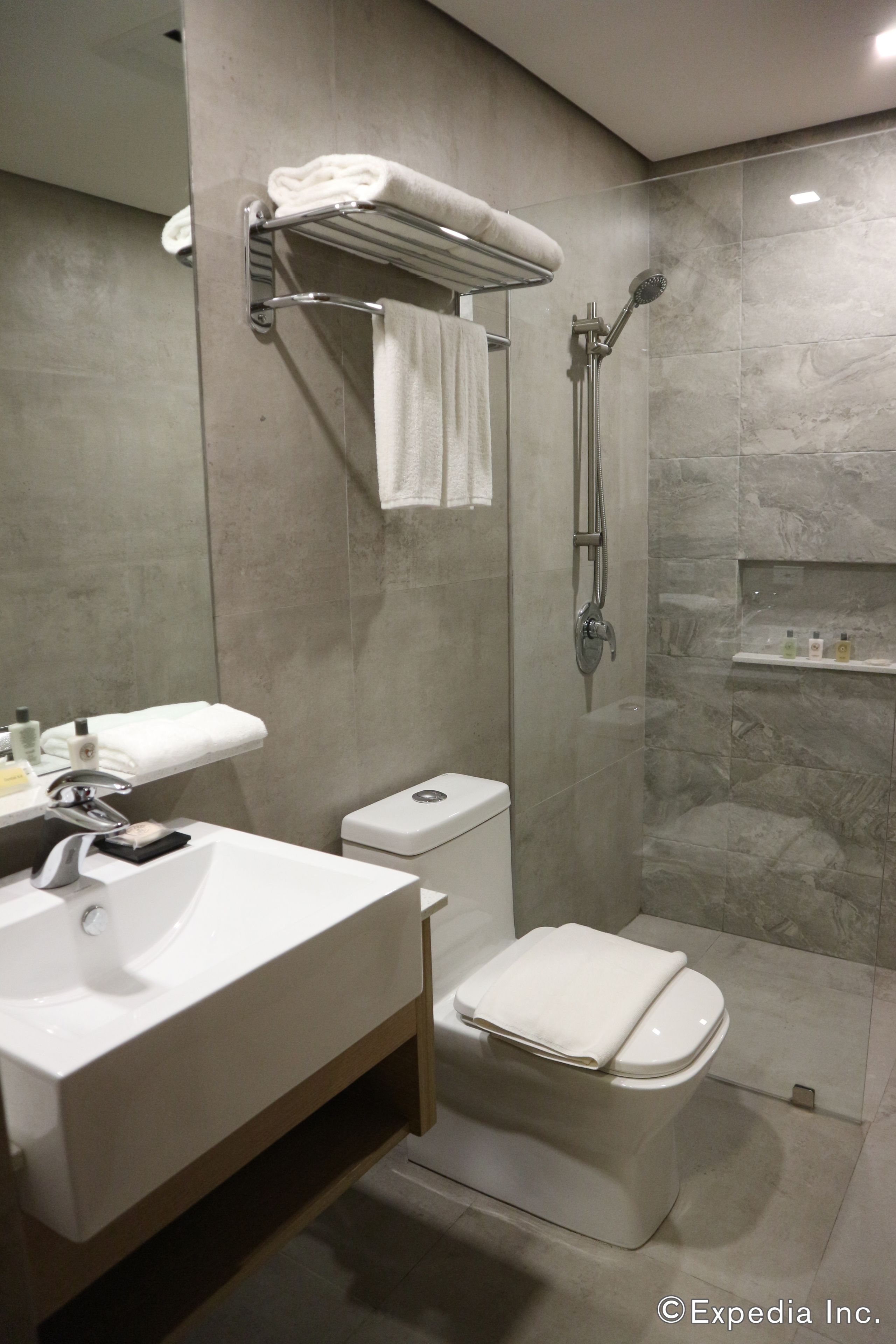 Une salle de bain moderne avec un lavabo, des toilettes et une douche, le tout de couleur blanche. On trouve un porte-serviettes au-dessus du lavabo et une étagère avec des articles de toilette.