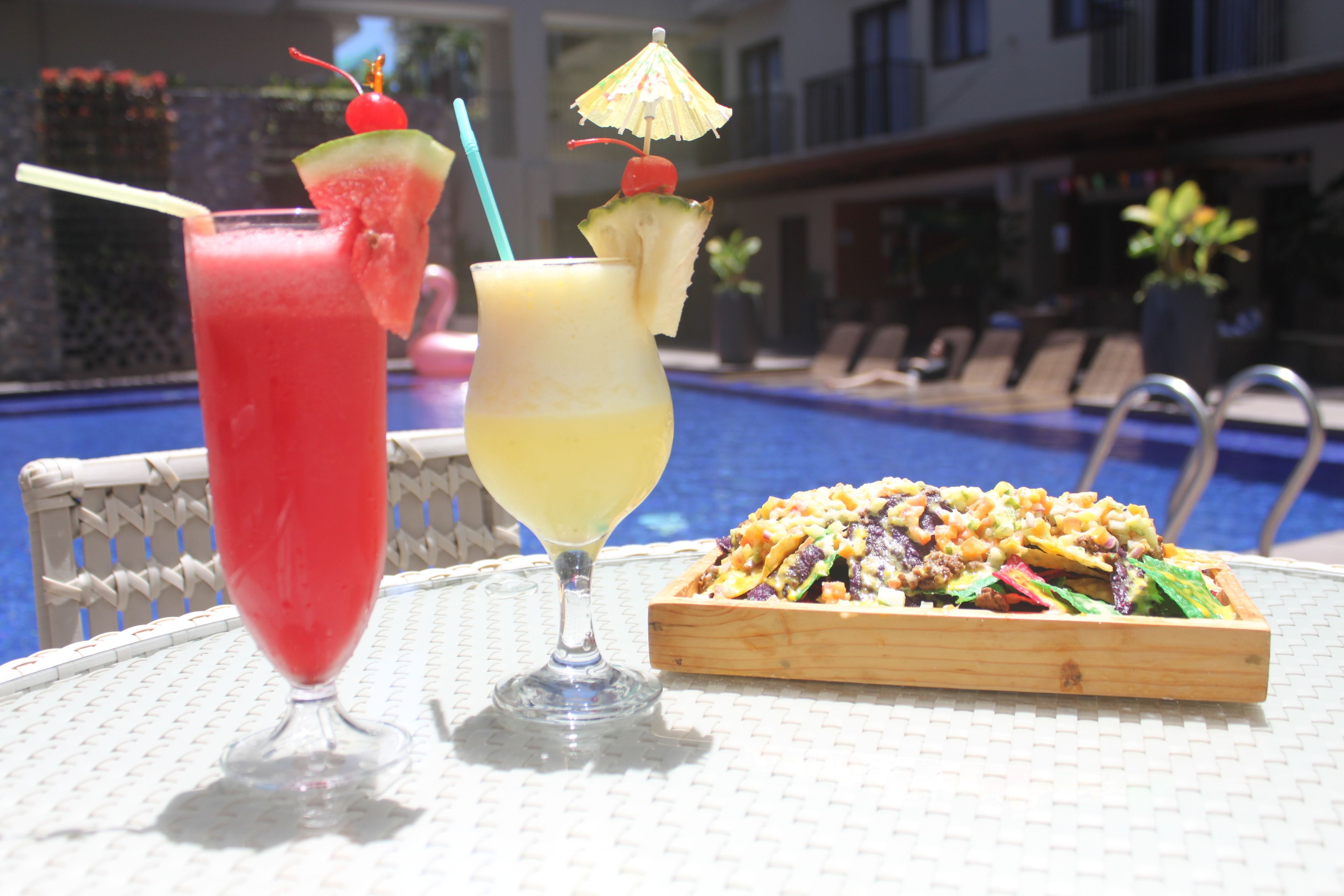 Deux boissons décorées de fruits et un plateau en bois contenant une salade, le tout posé sur une table au bord de la piscine.