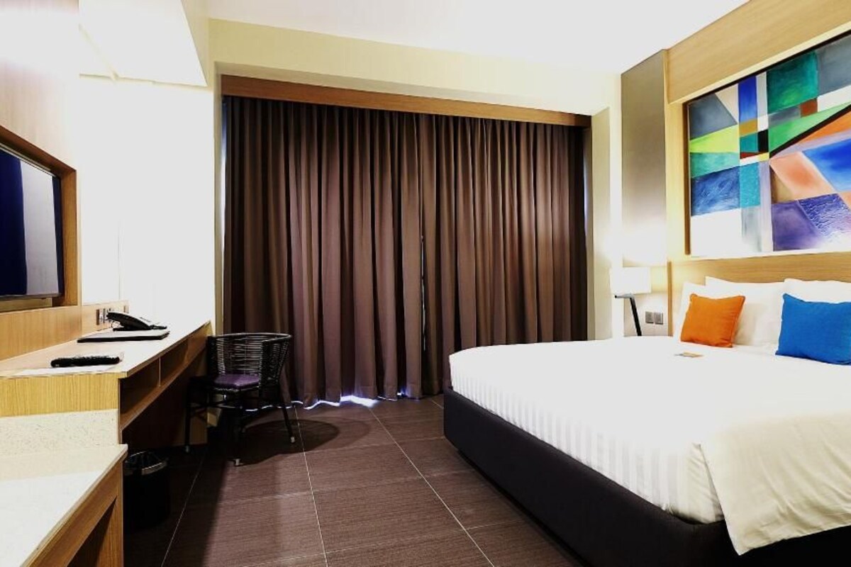 Habitación de hotel con cama, escritorio con computadora, televisor y ventana grande con cortinas.