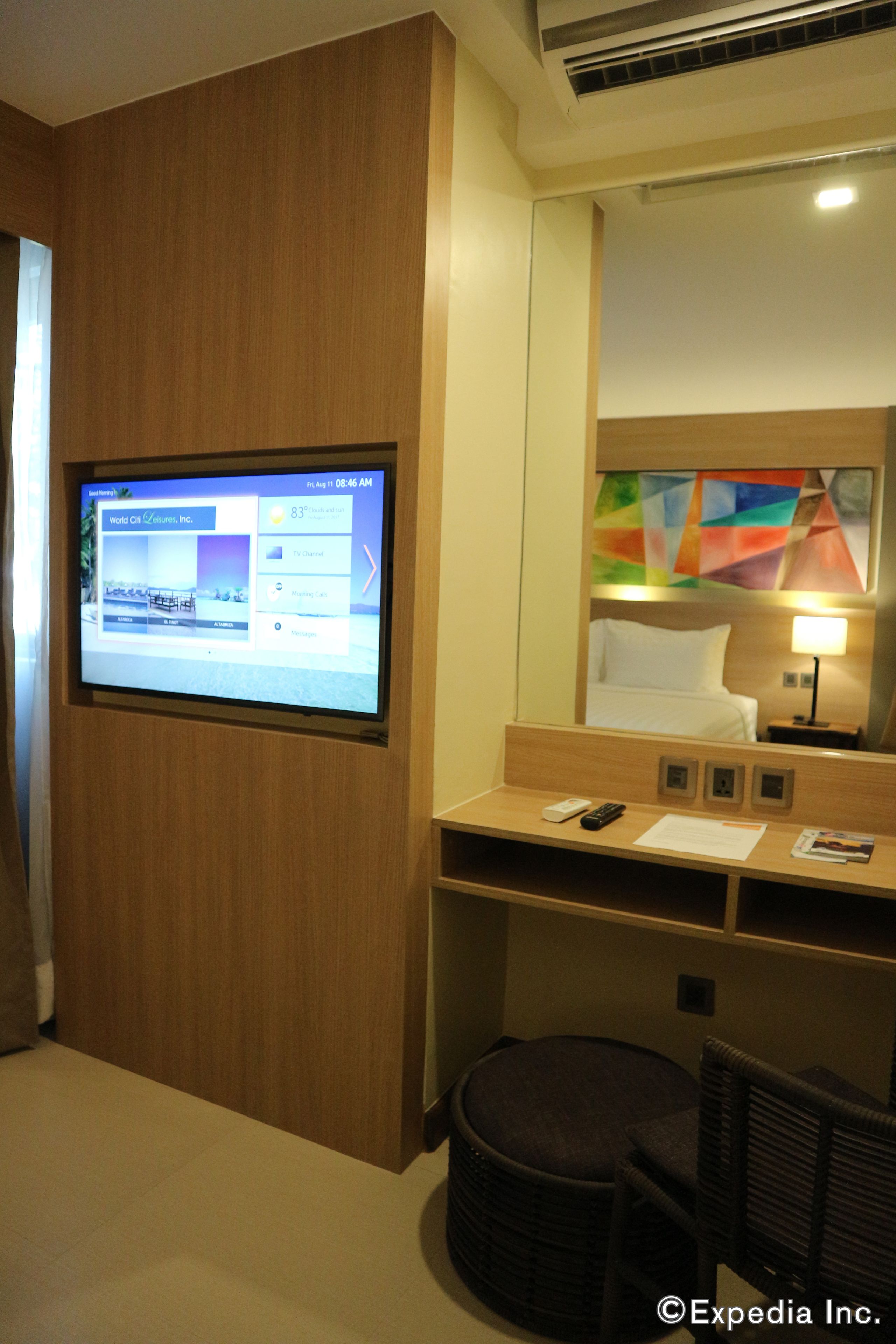 Habitación de hotel con una pantalla grande mostrando la página web del hotel, una cama, un escritorio con una lámpara, una silla y un otomanillo redondo.