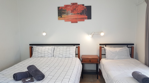Ein Zimmer mit zwei Betten, jedes mit einer eigenen gemusterten Tagesdecke. Dazwischen steht ein kleiner Nachttisch mit einer Lampe. Darüber befindet sich ein Wandbild, das eine rote Landschaft zeigt.