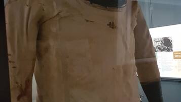 A mannequin in a hazmat suit with a gas mask, standing in a room with a glass wall and a poster in the background.