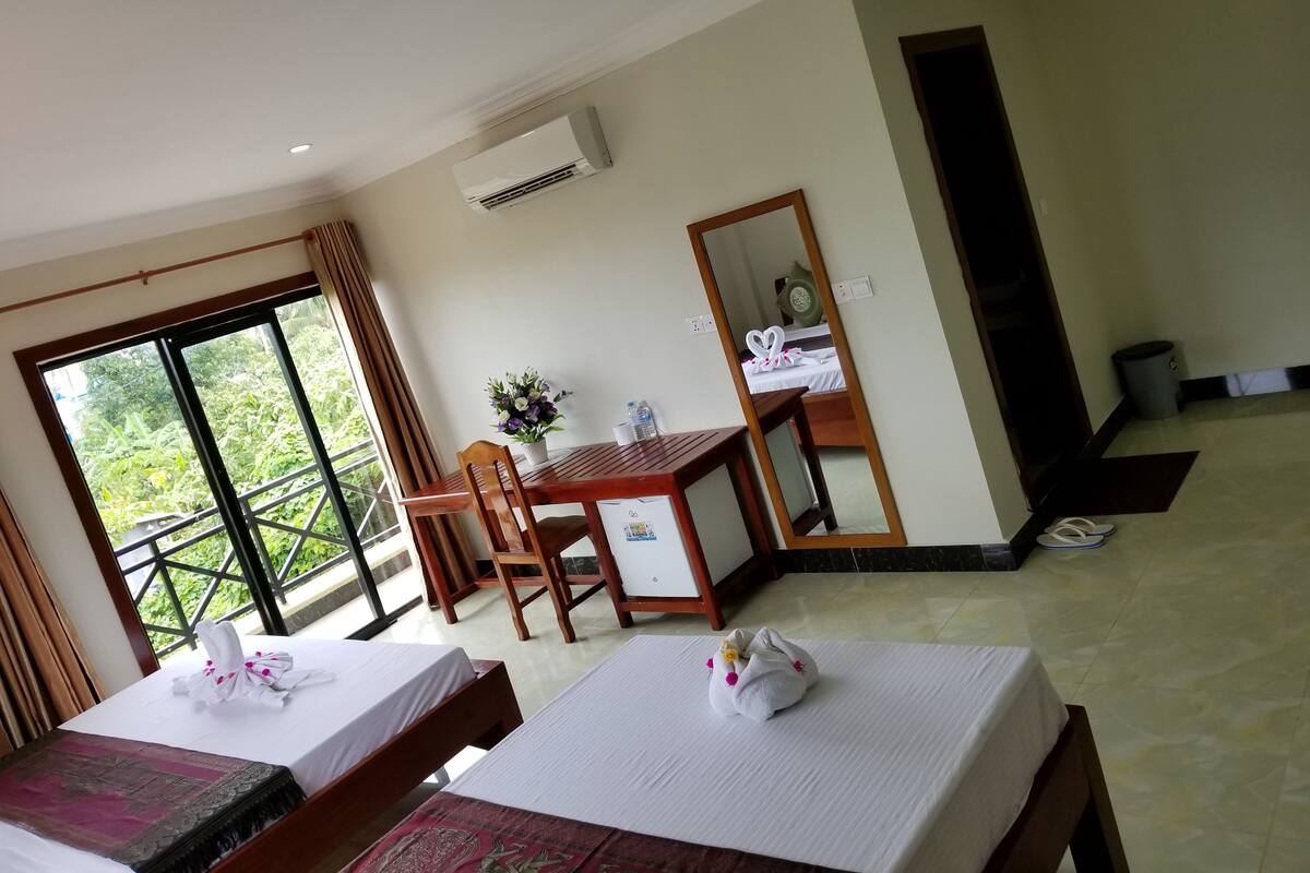 Quarto de hotel com duas camas, uma mesa de jantar, uma cadeira, um espelho e vista para vegetação pela janela.