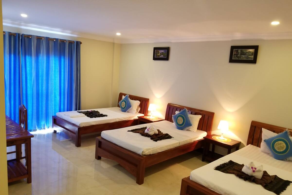 Quarto de hotel com três camas individuais, uma escrivaninha de madeira e uma cadeira.