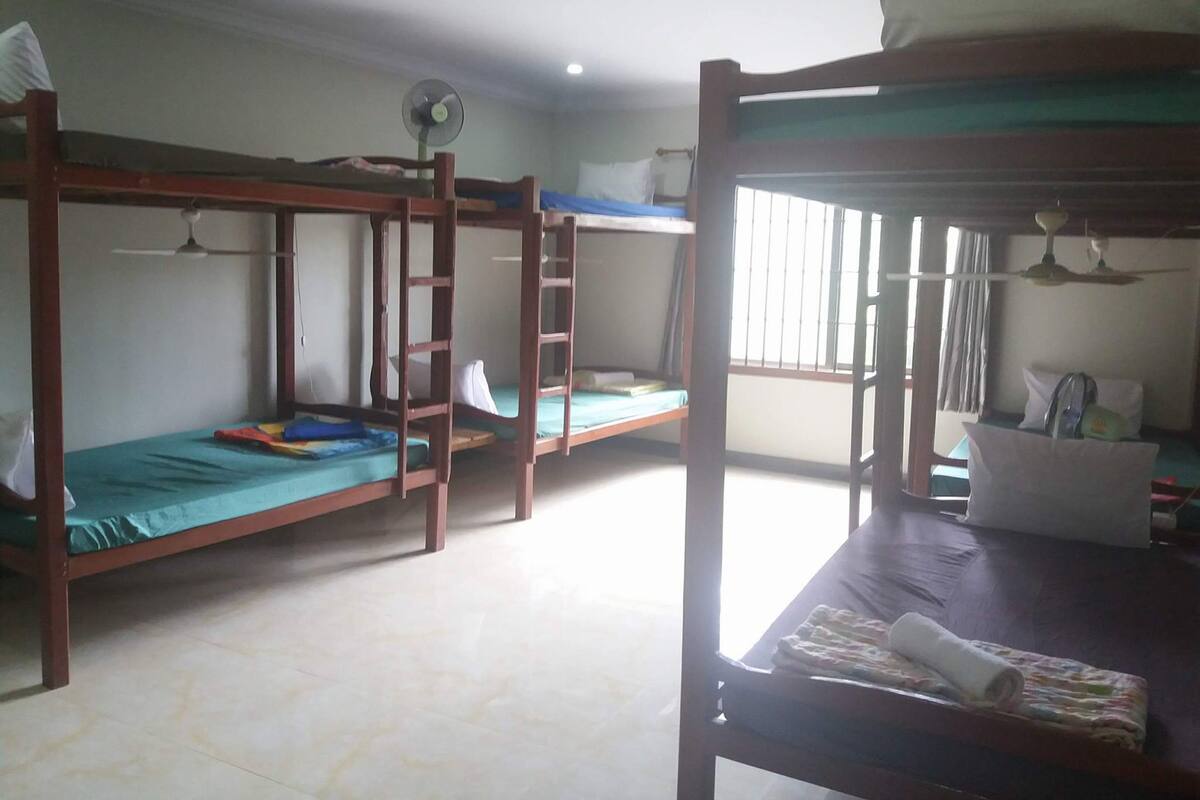 Quarto tipo dormitório com beliches, ventilador de teto e janela com grades.