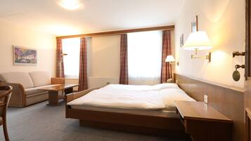 A hotel room with a bed, a sofa, a desk, and a chair. There are curtains on the windows, and a painting on the wall.