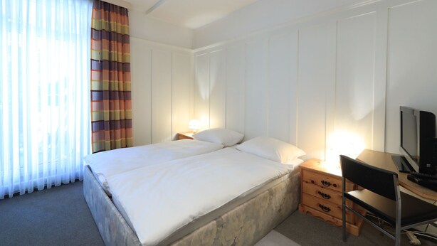 A hotel room with a bed, a desk, a chair, a TV, and a lamp. - Hotel Concordia Davos (Davos)