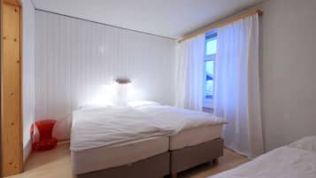 A bedroom with two beds, a red chair, and a window with white curtains.