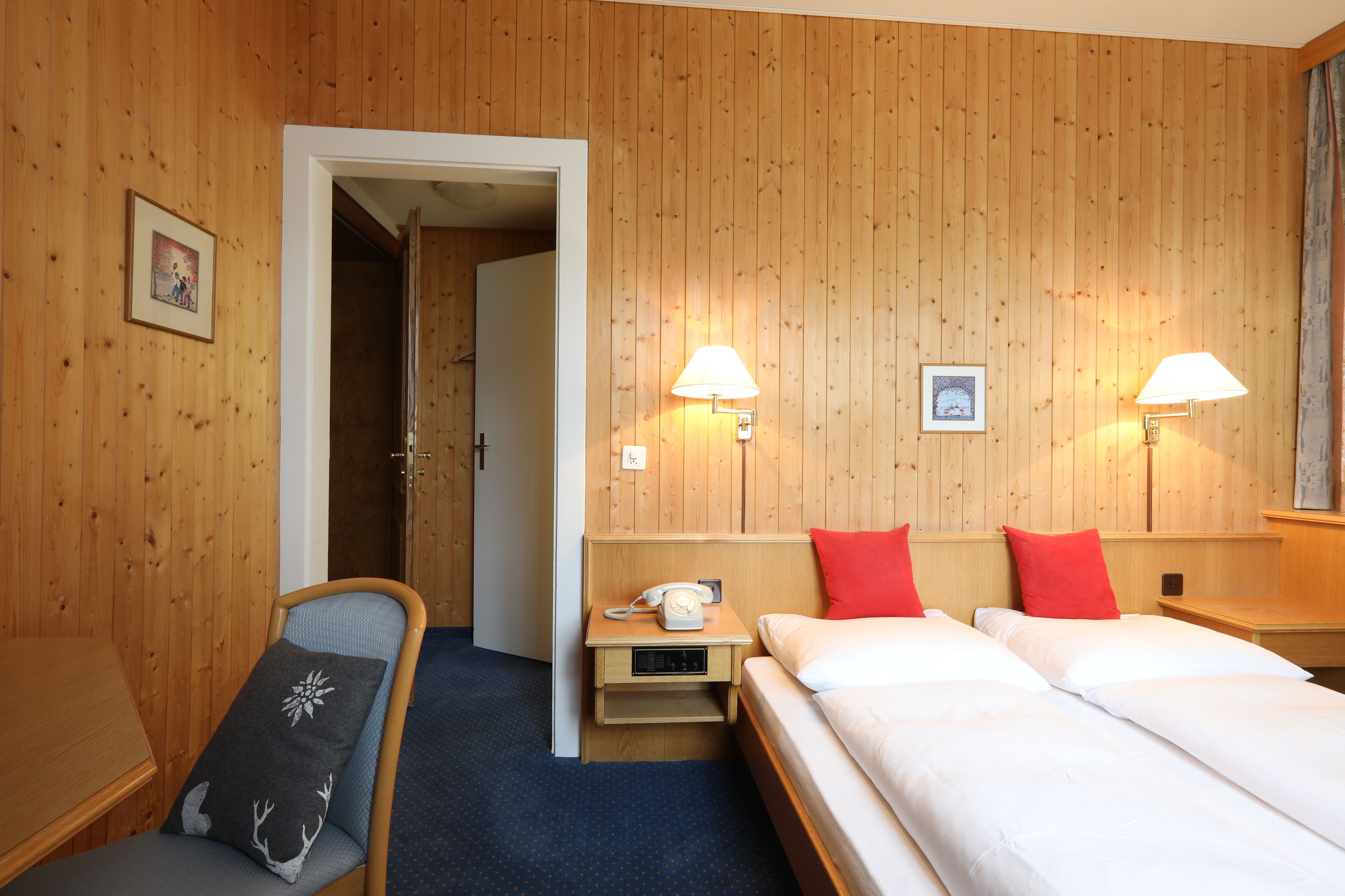 Habitación de hotel con paredes de paneles de madera, una cama con ropa blanca, dos lámparas de noche, una mesita de noche con un teléfono y una silla con cojín.