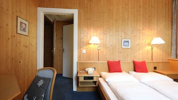 Habitación de hotel con paredes de paneles de madera, una cama con ropa blanca, dos lámparas de noche, una mesita de noche con un teléfono y una silla con cojín.