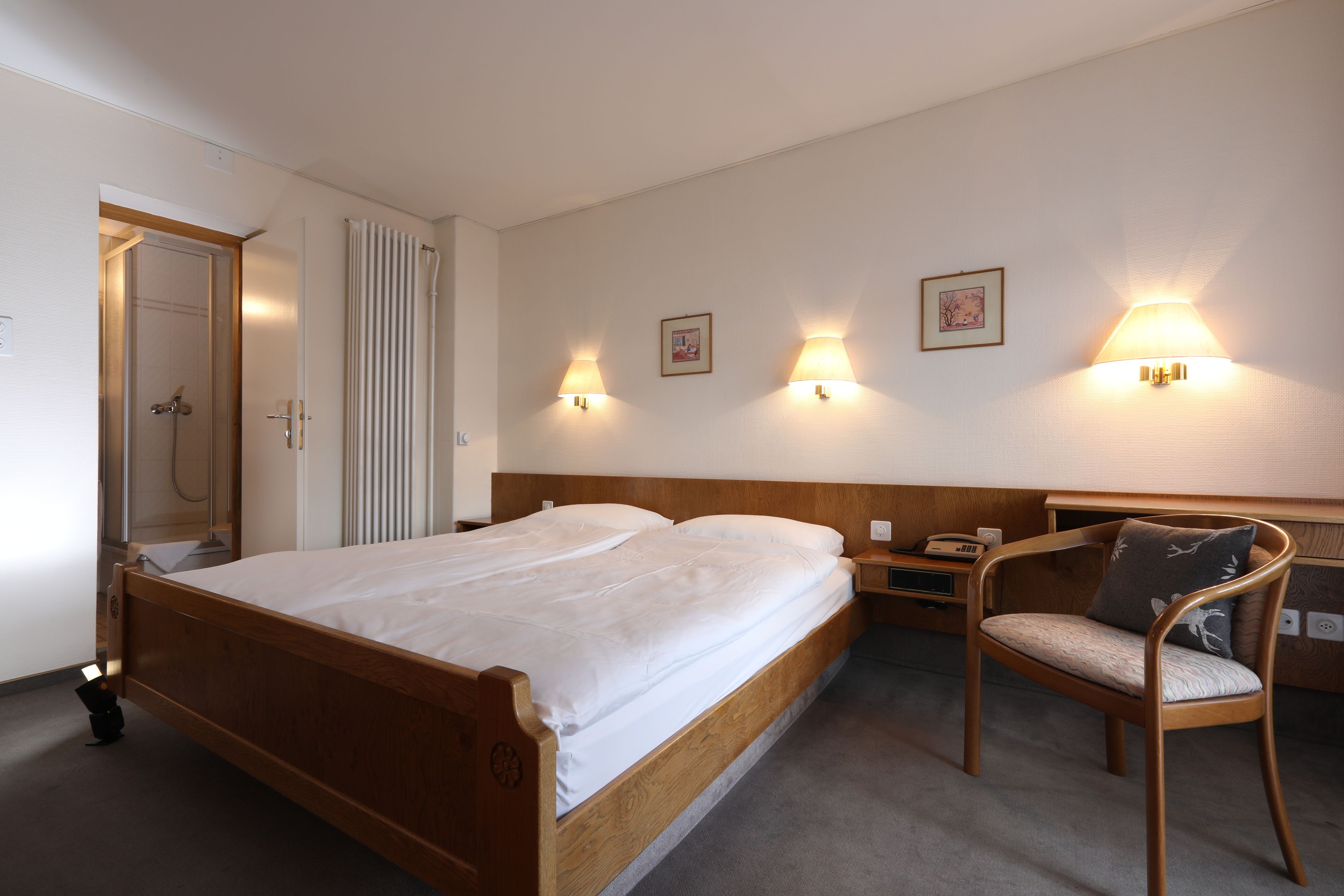 A hotel room with a wooden bed, a chair with a cushion, two wall-mounted lamps, and a framed picture on the wall.