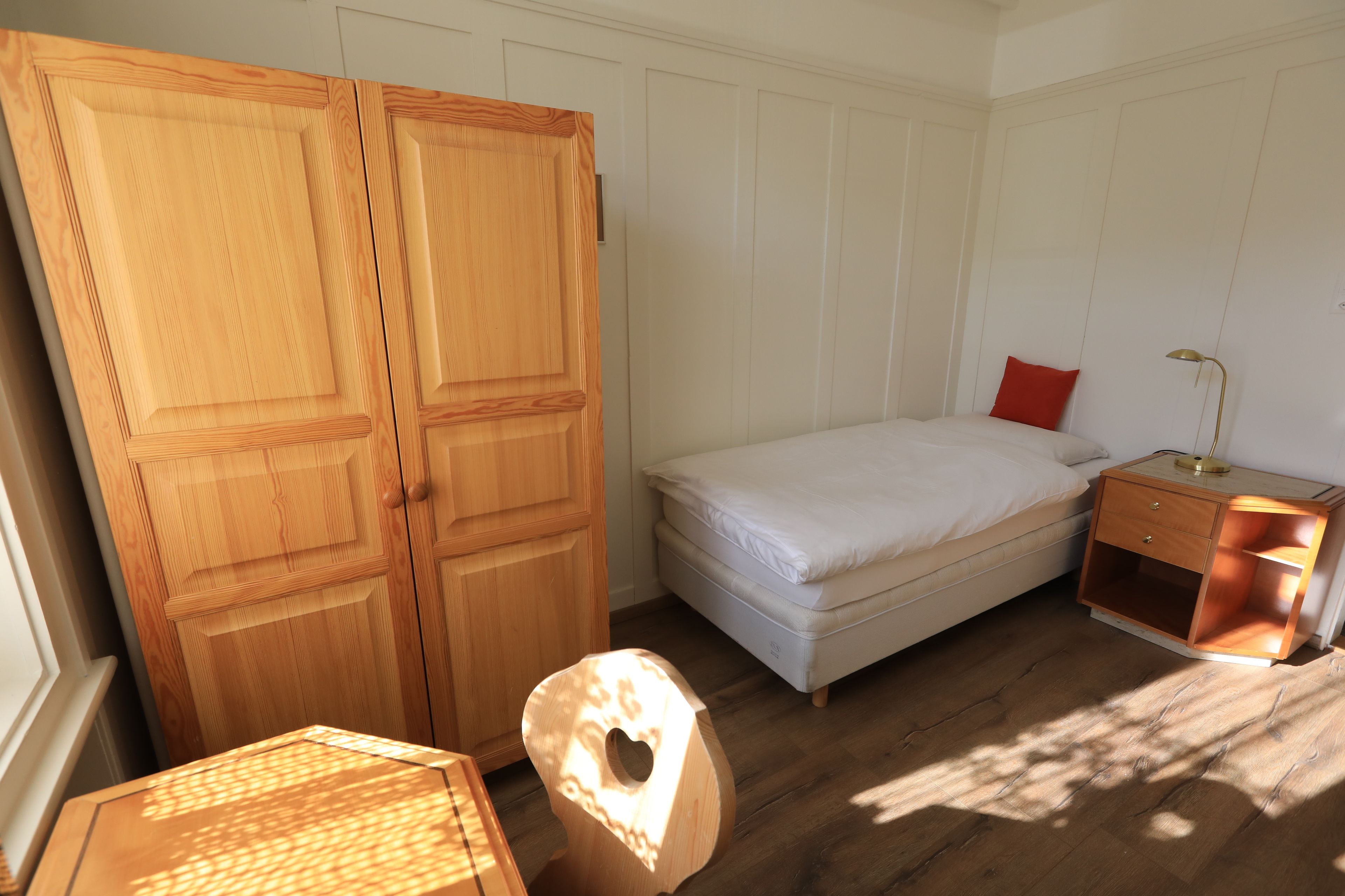 Un dormitorio con cama, mesita de noche, armario de madera y una pequeña mesa.