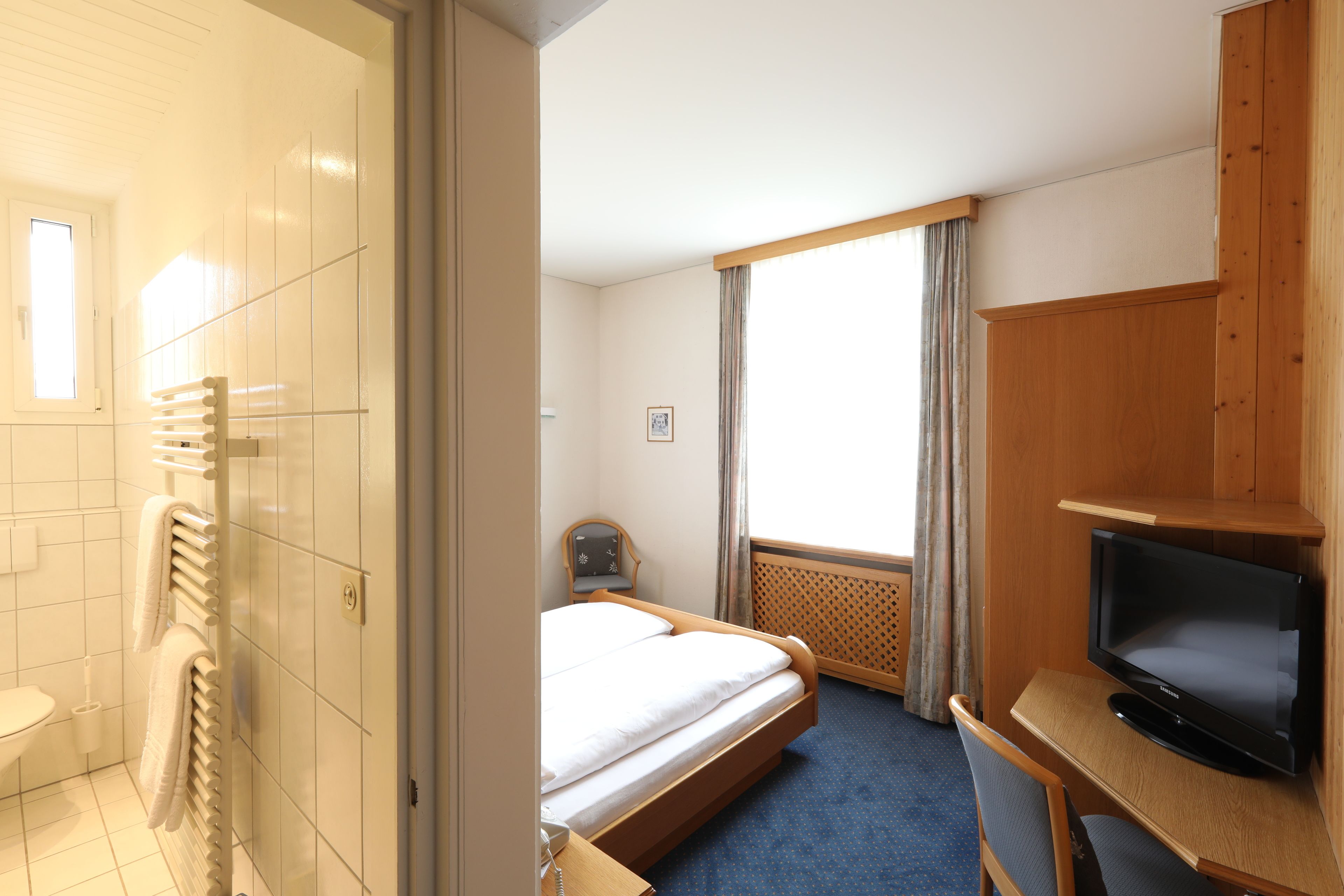 A hotel room with a bed, a desk with a television, a window with curtains, and a bathroom with a towel rack.