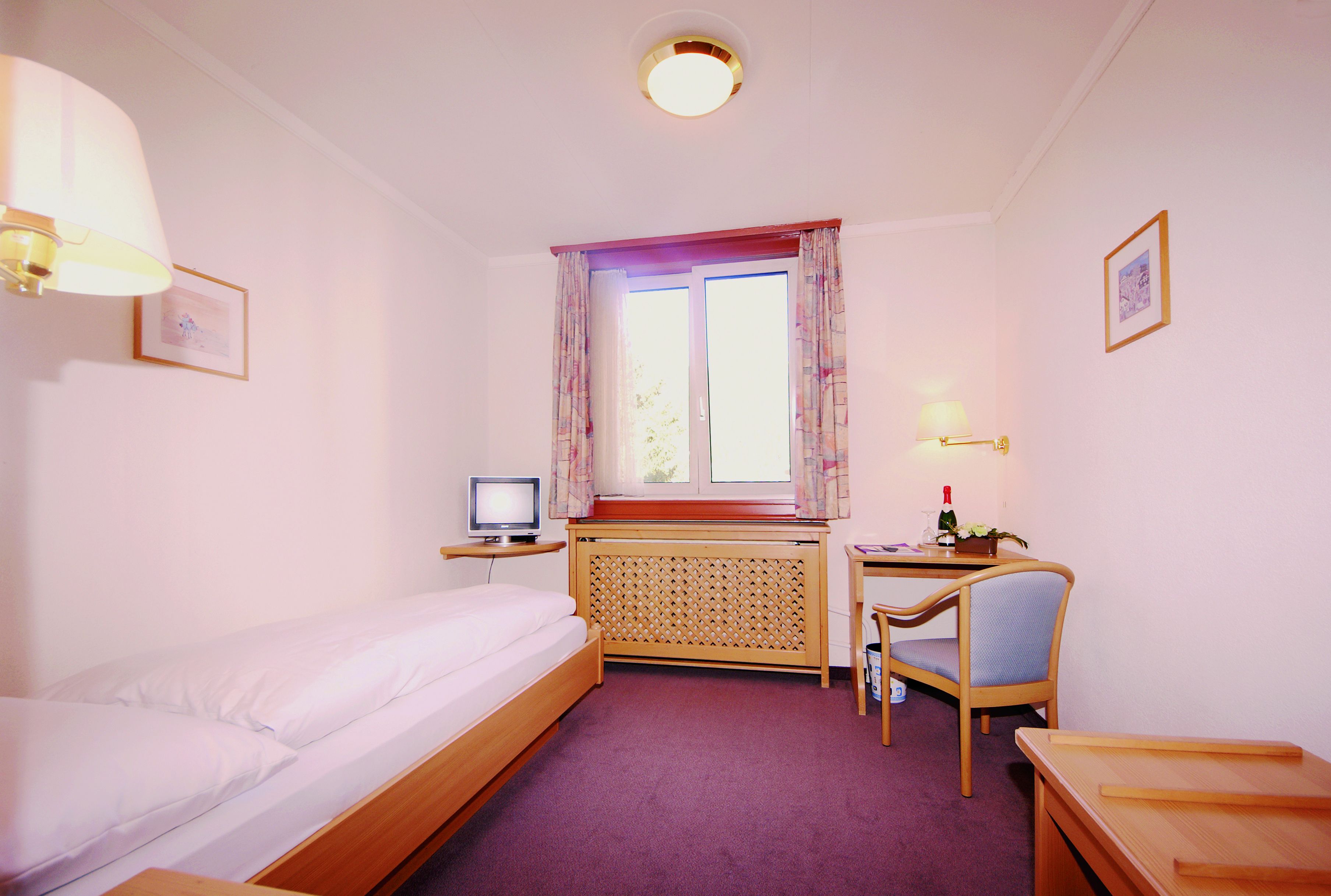 A bedroom with a bed, a desk, a chair, a television, and a window with curtains.