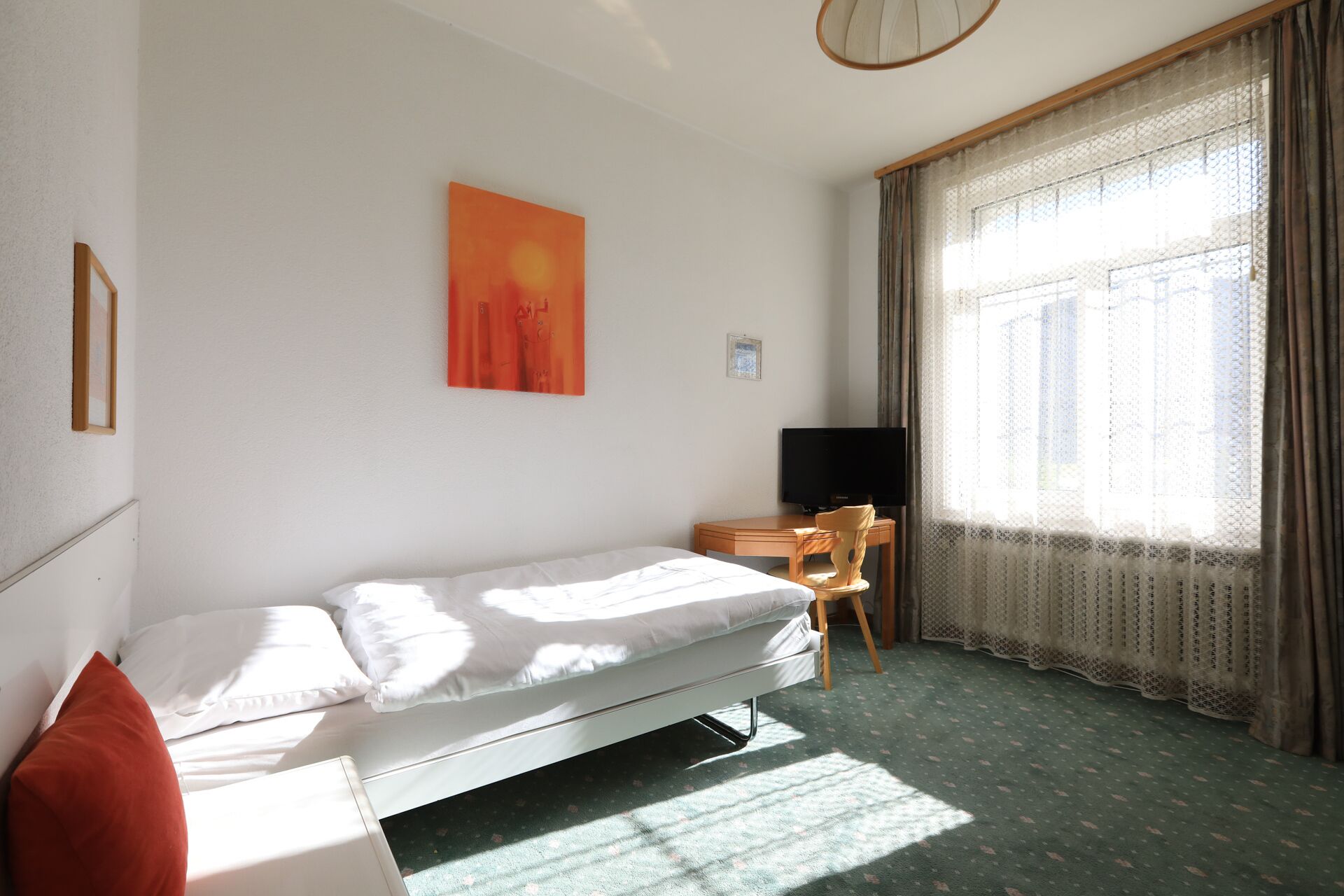 Una habitación pequeña y luminosa con una cama individual, un escritorio con una silla, un televisor y una ventana con cortinas.