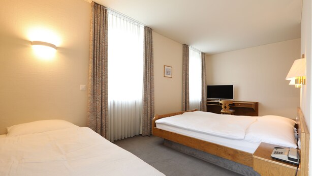 A hotel room with two single beds, a flat-screen TV, a bedside table with a lamp, and a window with curtains. - Hotel Concordia Davos (Davos)