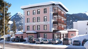 A multi-story hotel with a snow-covered roof, a visible balcony on the second floor, and a parking lot with several cars. - Hotel Concordia Davos (Davos)