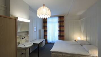 A hotel room with a bed, bedside table, lamp, desk, chair, and a large window with curtains.