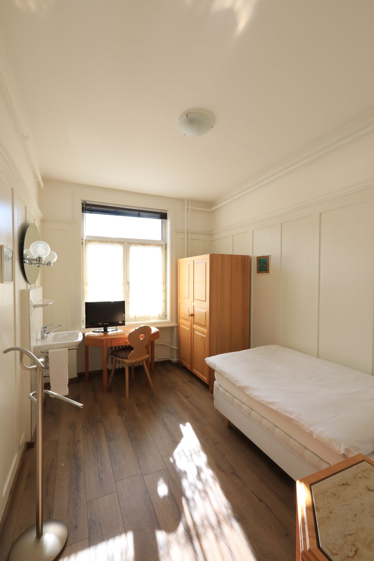 Una habitación pequeña y luminosa con piso de madera, una cama, un escritorio con silla, un televisor y un guardarropa.