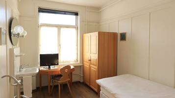 Una habitación pequeña y luminosa con piso de madera, una cama, un escritorio con silla, un televisor y un guardarropa.