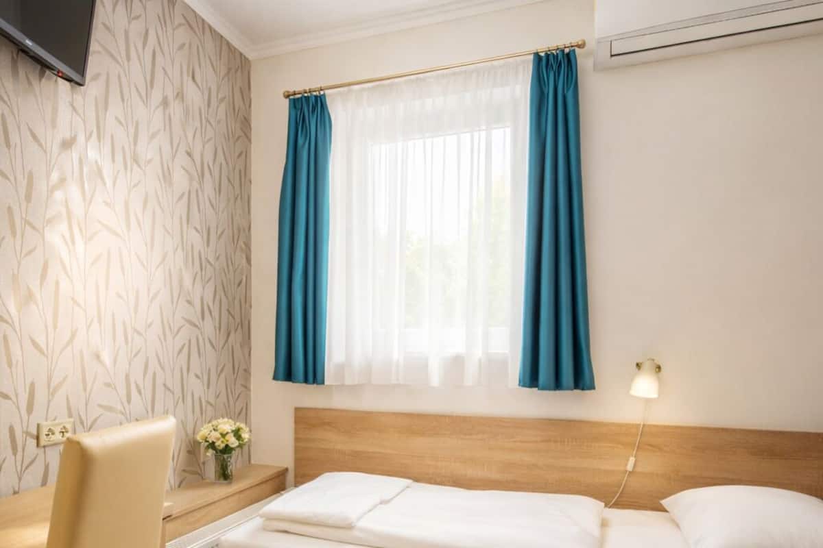 A hotel room with a bed, a desk, a chair, a window with curtains, a wall-mounted TV, and a radiator.