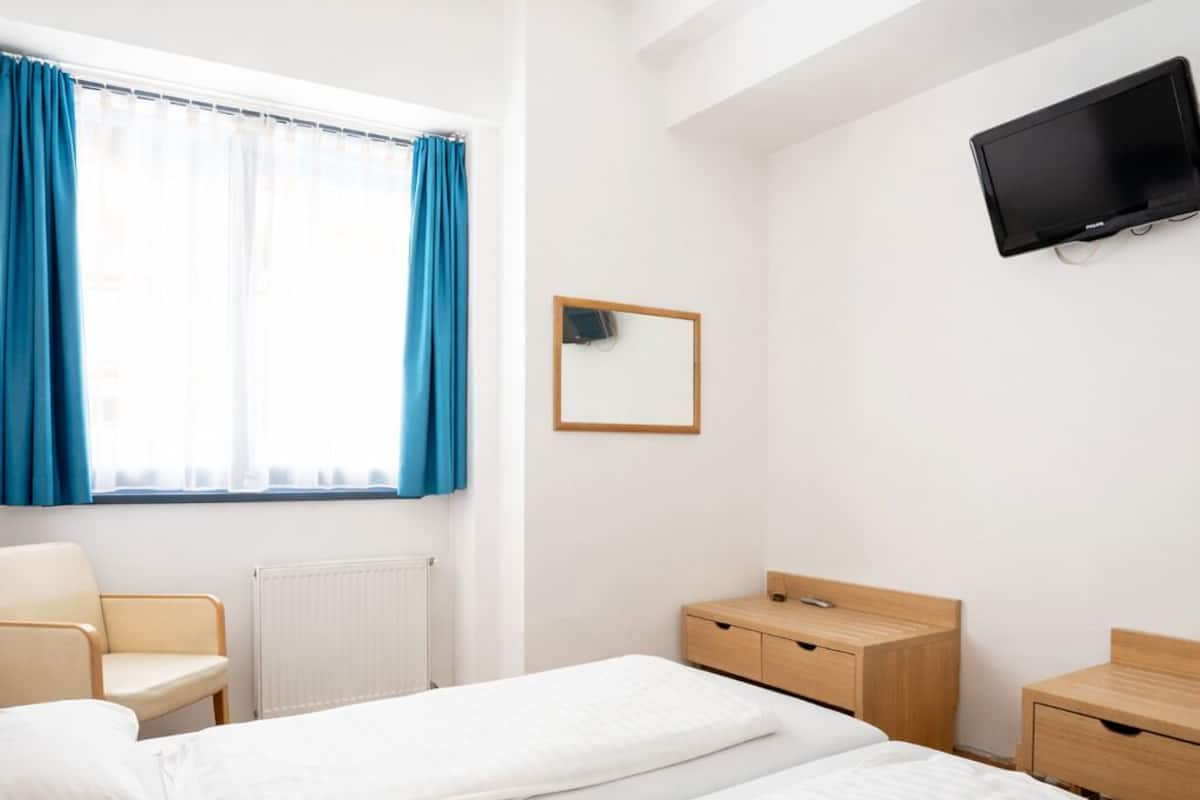 A hotel room with two single beds, a wooden dresser, a chair, a TV mounted on the wall, and a window with blue curtains.