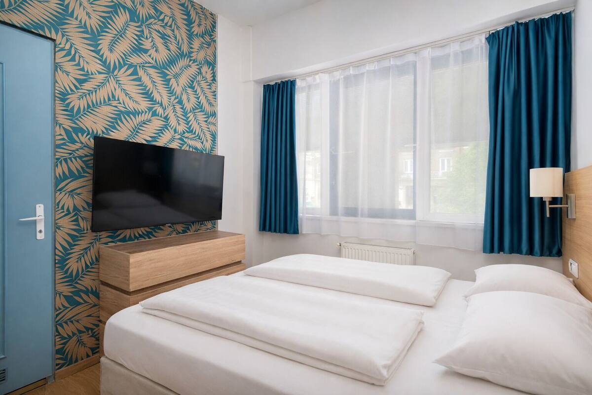 A hotel room with a flat-screen TV on a wooden dresser, two beds with white linens, a window with blue curtains, and a wall with a leaf-pattern wallpaper.