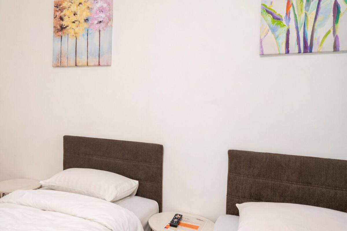 A hotel room with two beds, a small round table with a remote control, and two paintings on the walls.