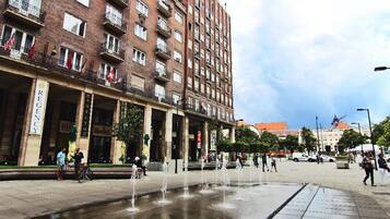 Una plaza pública con una fuente, un edificio de varios pisos y balcones, y un cielo despejado.