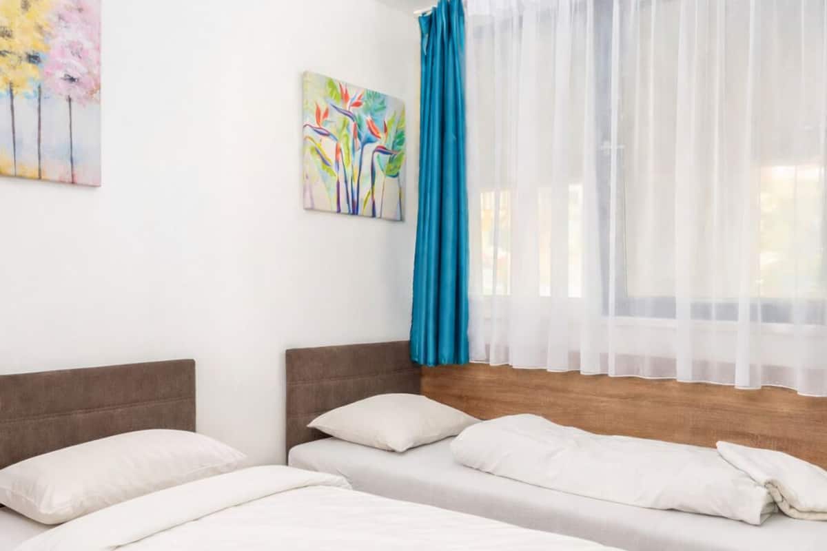 A room with two beds, white bedding, a wooden headboard, a blue curtain, and a painting on the wall.