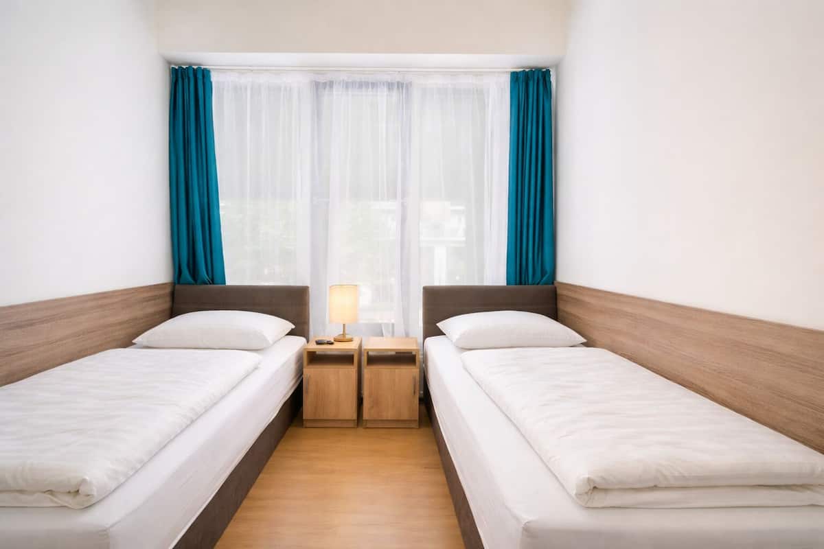 A twin room with two single beds, a nightstand with a lamp, and a window with sheer curtains.