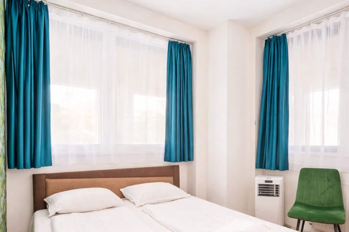 A neatly made bed in a room with two windows, each with blue curtains.