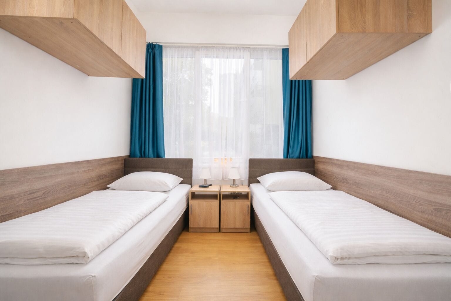 A twin room with two single beds, wooden headboards, a small bedside table with a lamp, and a window with blue curtains.