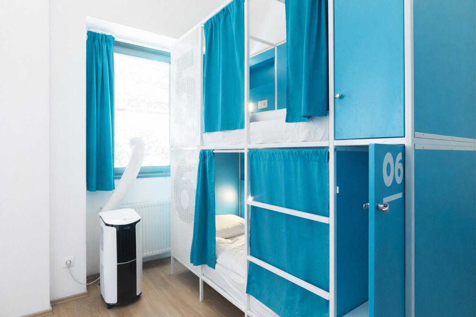 A dormitory room with bunk beds, a window with curtains, a heater, and a trash bin.