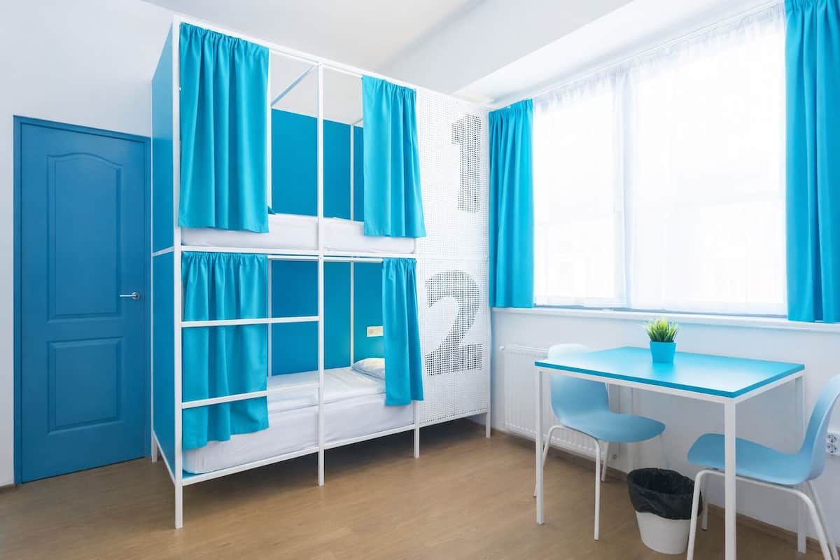A room with bunk beds, a desk, chairs, and a window with curtains.