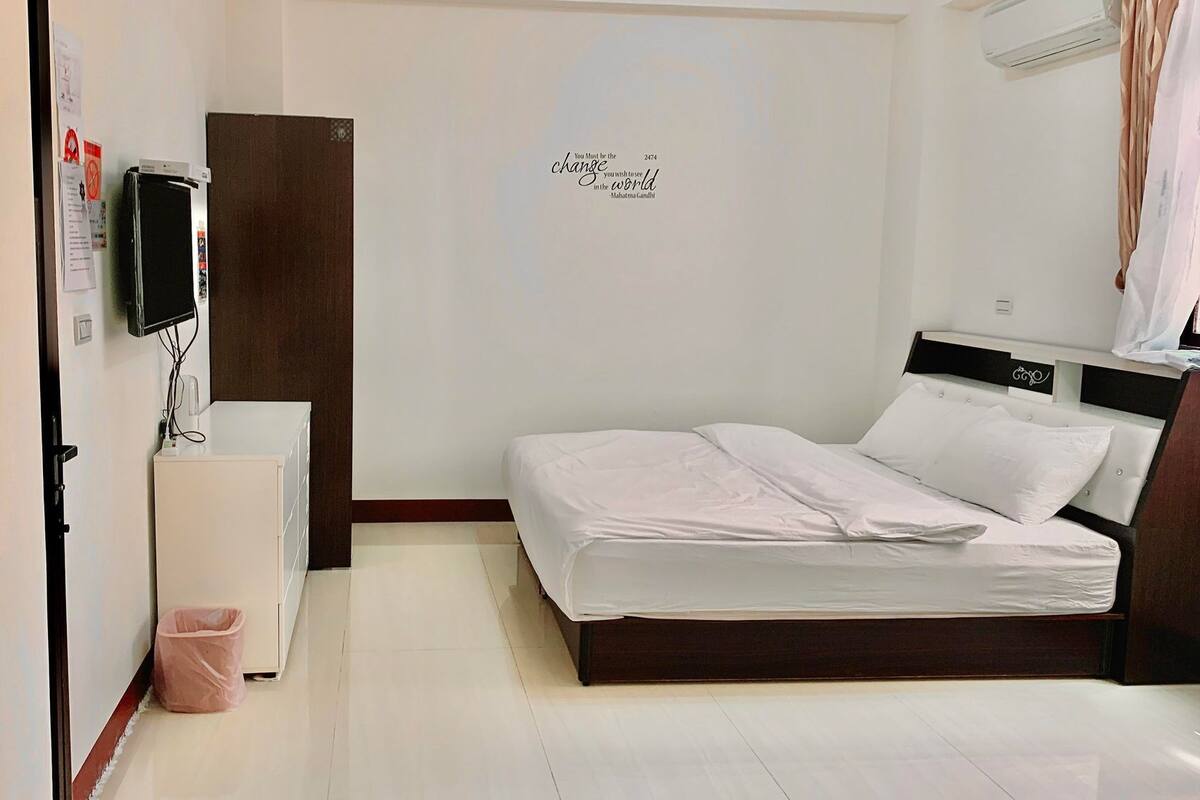 A bedroom with a bed, a TV, a small cabinet, and a wall with a motivational quote.