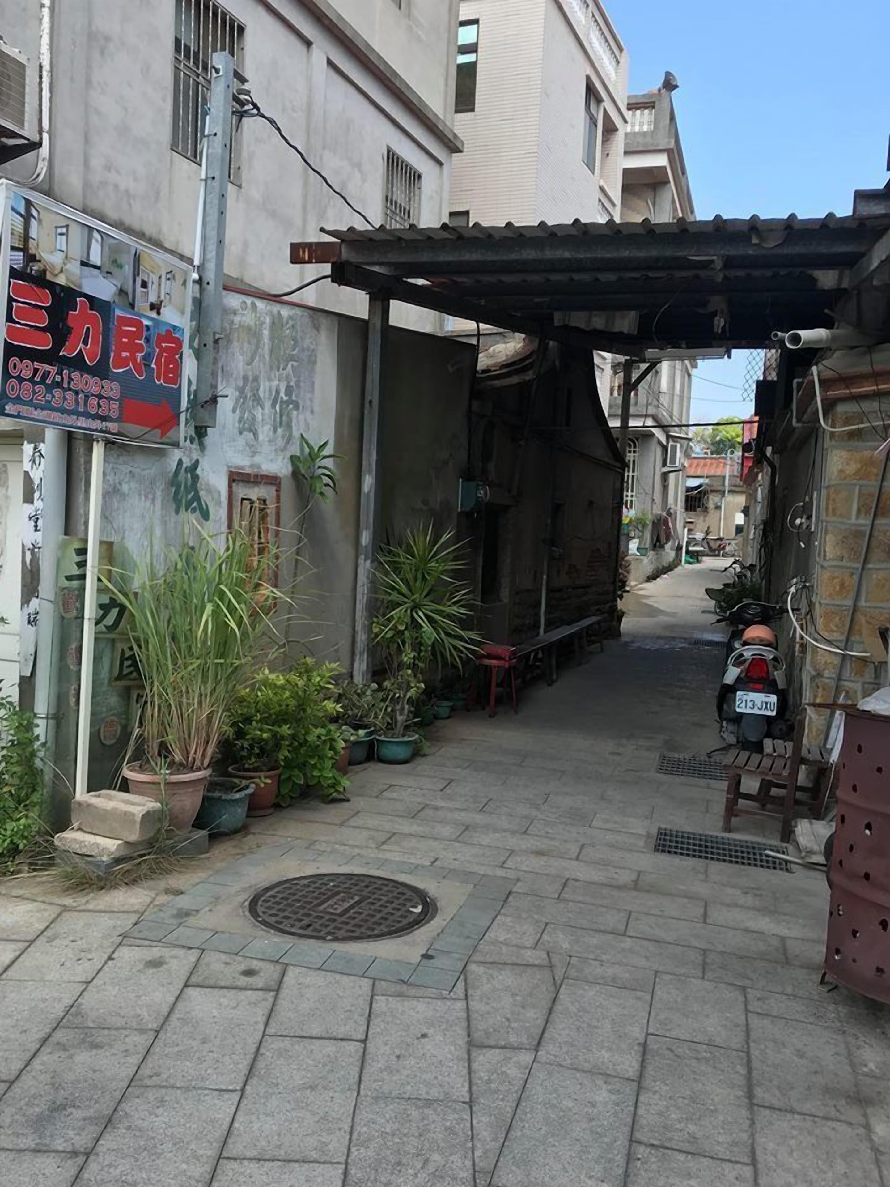 A narrow alleyway with buildings on either side, potted plants, and a motorcycle parked on the right.