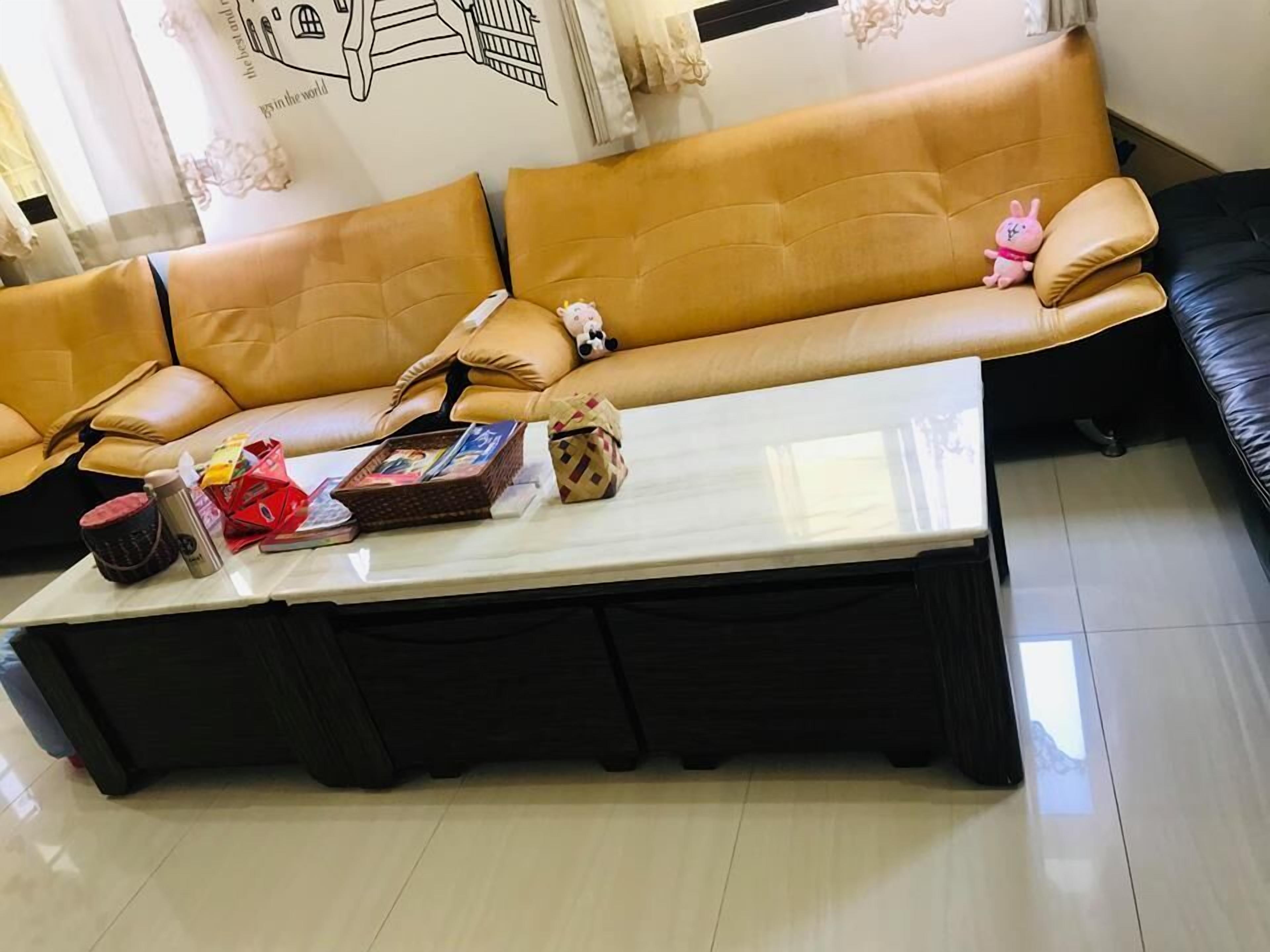 Una sala de estar con un sofá de cuero marrón, una mesa de centro blanca con varios objetos, y un sofá negro al fondo.