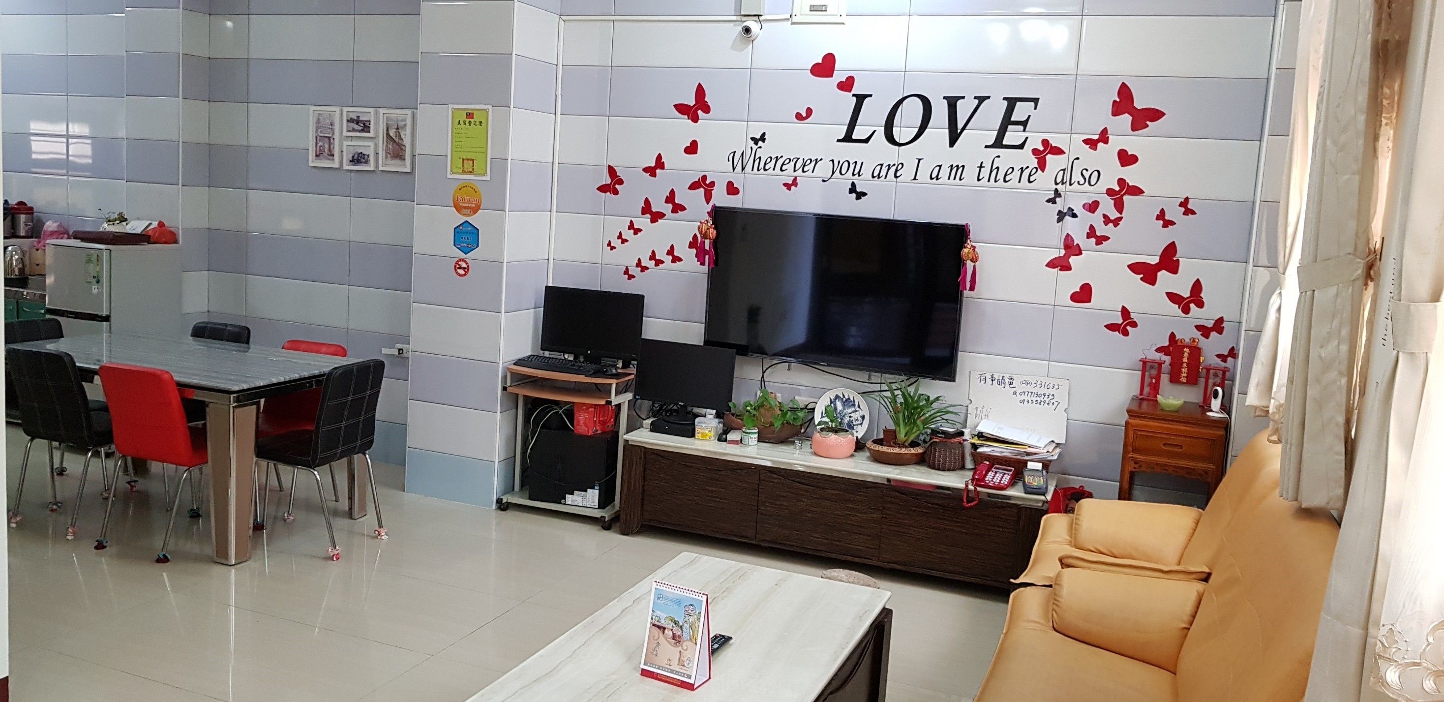 Una sala de estar moderna con un sofá, una mesa de centro y un televisor. Cuenta con una zona de comedor con una mesa y sillas. La habitación presenta una pared con un cartel que dice 'LOVE' y mariposas.