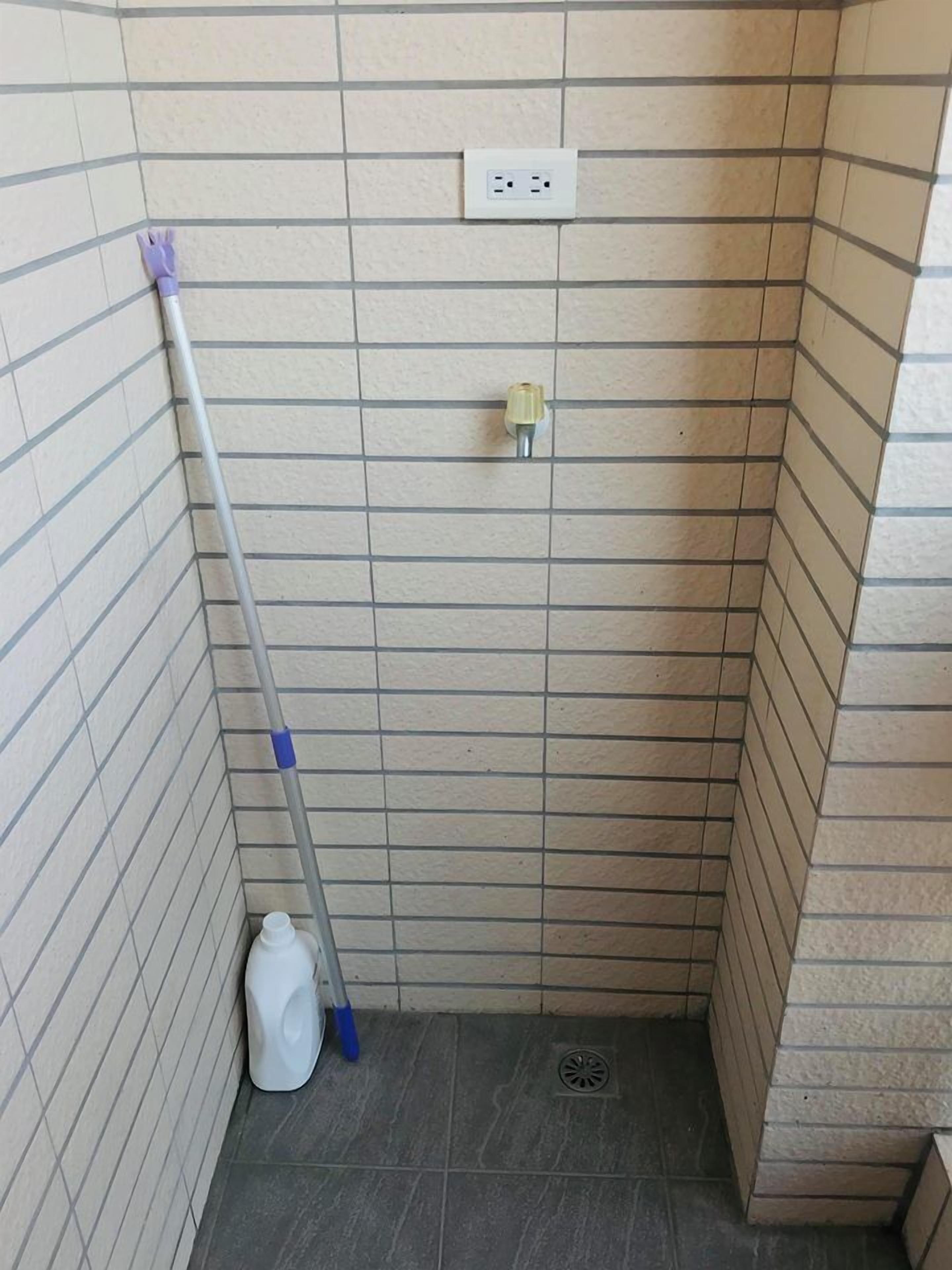 A small, tiled bathroom with a shower area, a mop leaning against the wall, and a white bottle on the floor.
