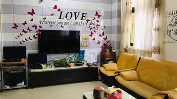 A living room with a large flat-screen TV, a yellow sofa, a coffee table with books and a thermos, and a wall with a 'LOVE' sign and butterflies.