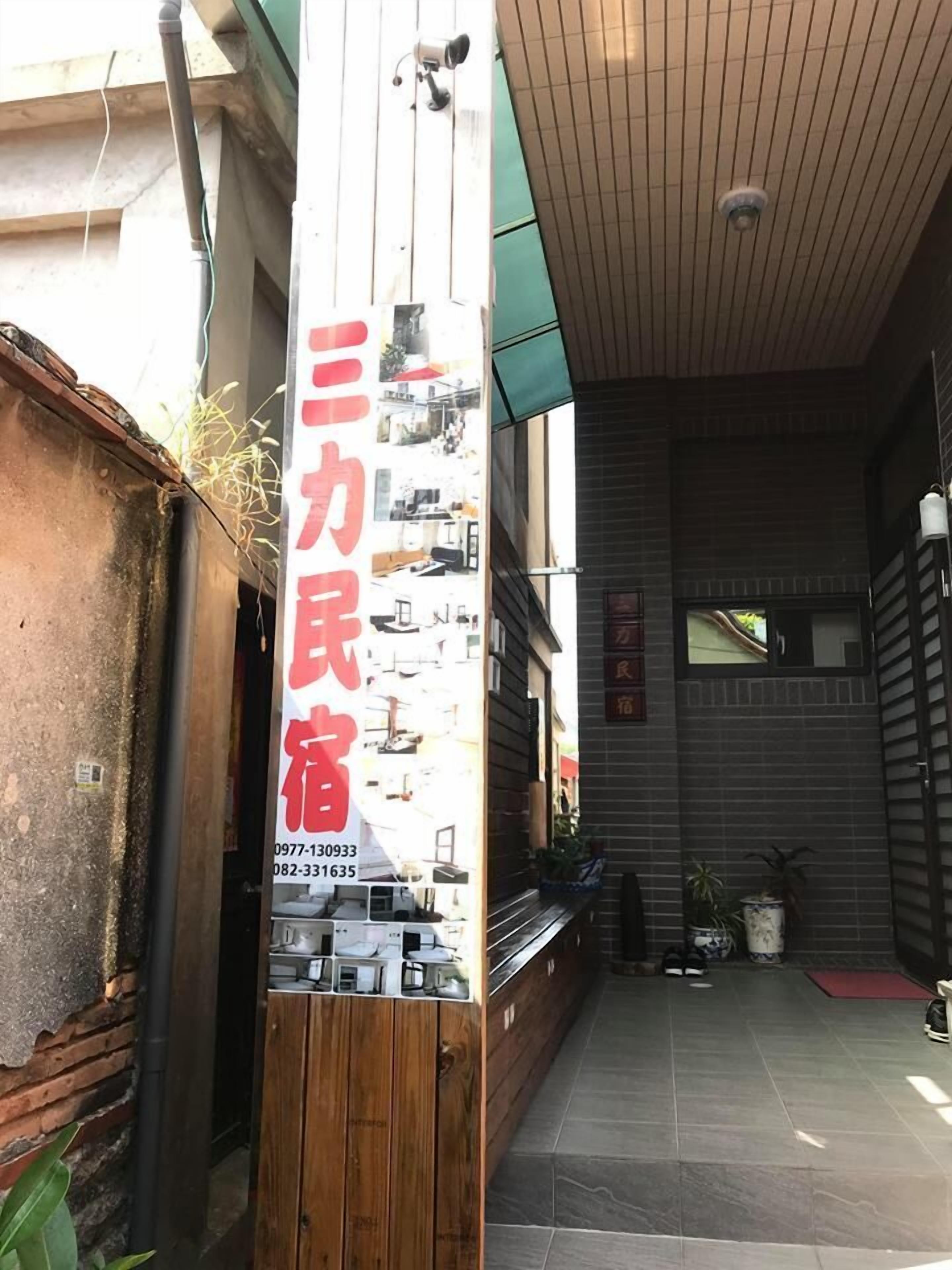 A narrow alleyway with a wooden fence on one side and a building entrance on the other. A vertical sign with red text is visible, indicating a business or service.